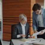 Two business colleagues discussing reports in modern office indoors. Businesswoman sitting at the table talking his male partner